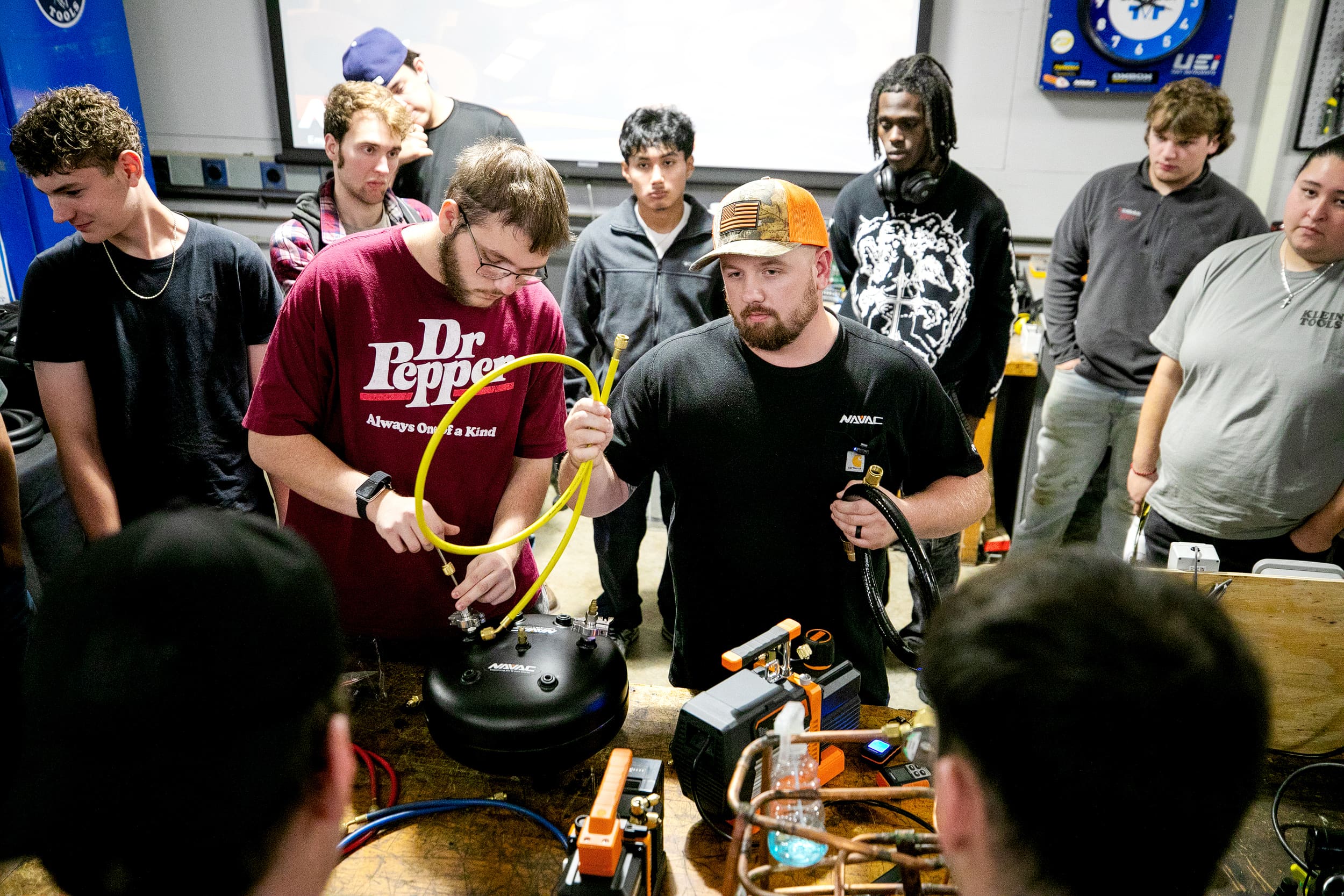 A group of students gather around two instructors demonstrating HVAC equipment and tools on a workbench in a classroom setting.