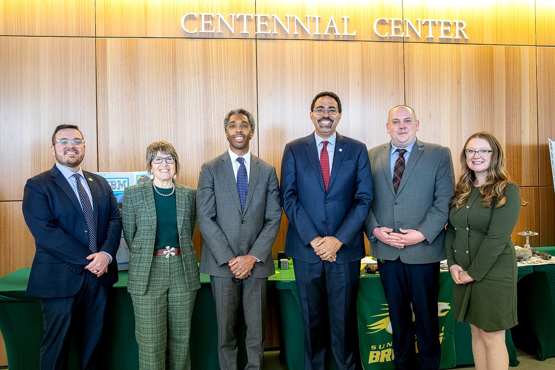 Six professionally dressed people stand in a row, smiling, in front of a wooden wall labeled "Centennial Center" with tables and green tablecloths behind them.