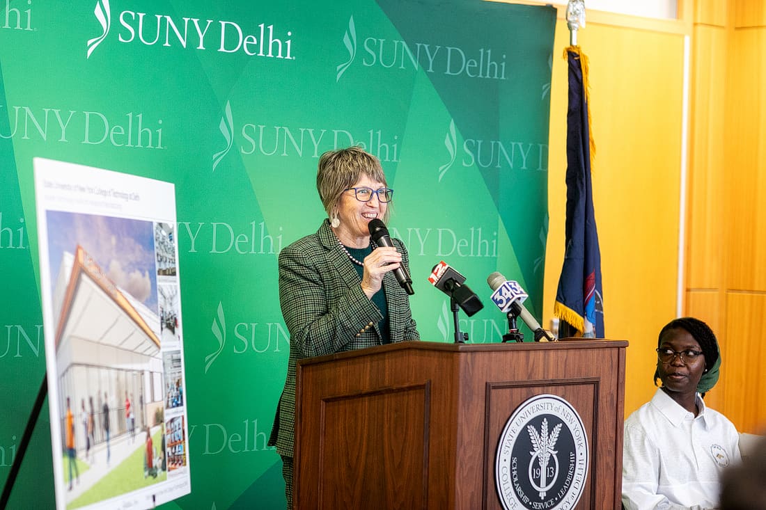A woman speaks at a podium with microphones in front of a SUNY Delhi backdrop; another person sits nearby. An architectural rendering is displayed on an easel.