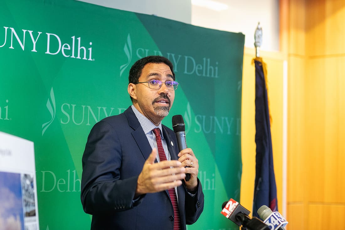 A man in a suit speaks into a microphone at a podium with news microphones, standing in front of a SUNY Delhi backdrop.
