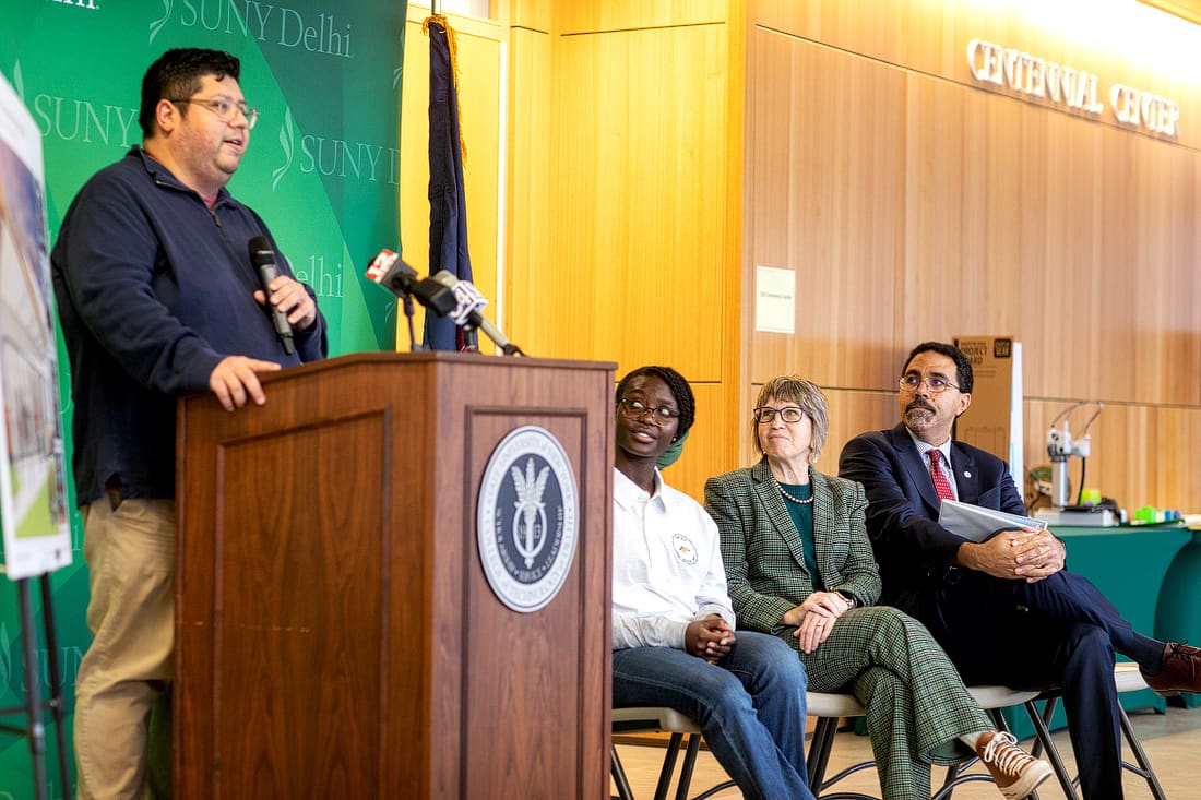 A man speaks at a podium with three people seated beside him, in front of a SUNY Delhi backdrop in a brightly lit room.