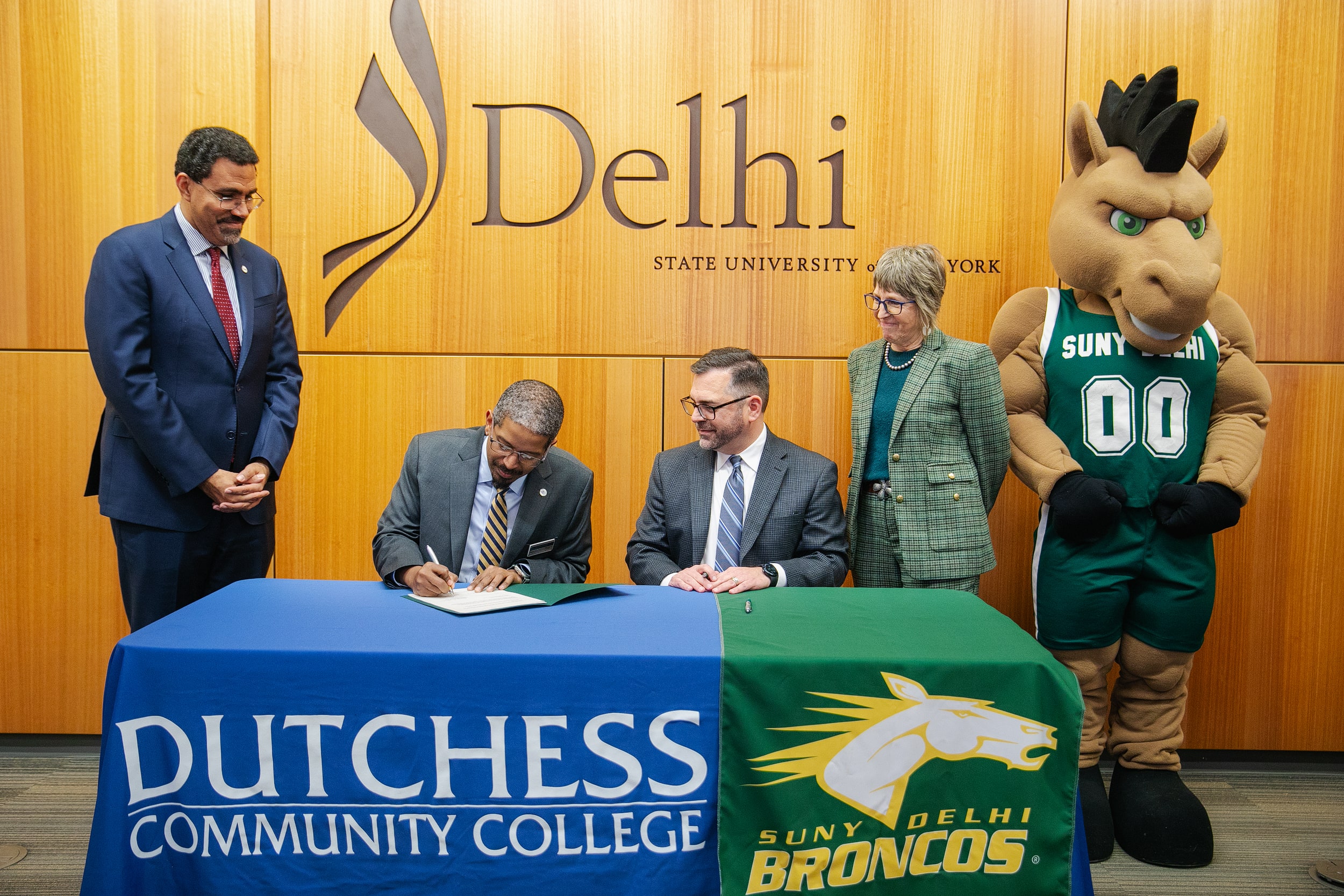 Four people, including two men in suits and one woman, sit and stand at a table with Dutchess Community College and SUNY Delhi logos; a mascot in a Broncos jersey stands to the right.