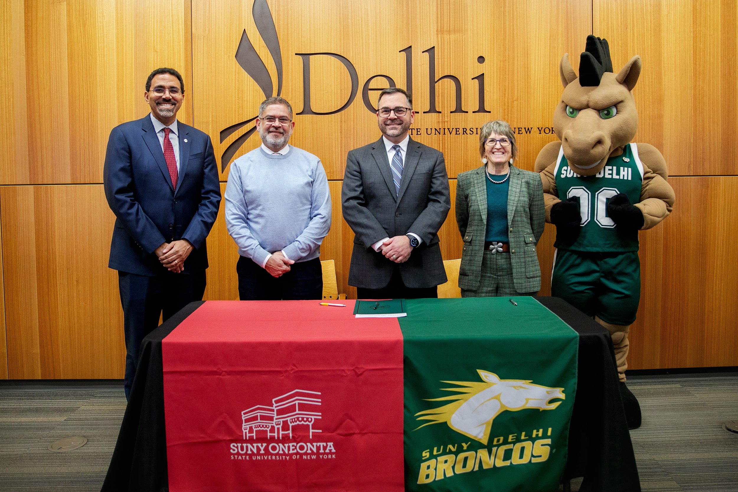 Five people, including a mascot, stand behind a table with SUNY Oneonta and SUNY Delhi banners in front of a wood wall with the SUNY Delhi logo.