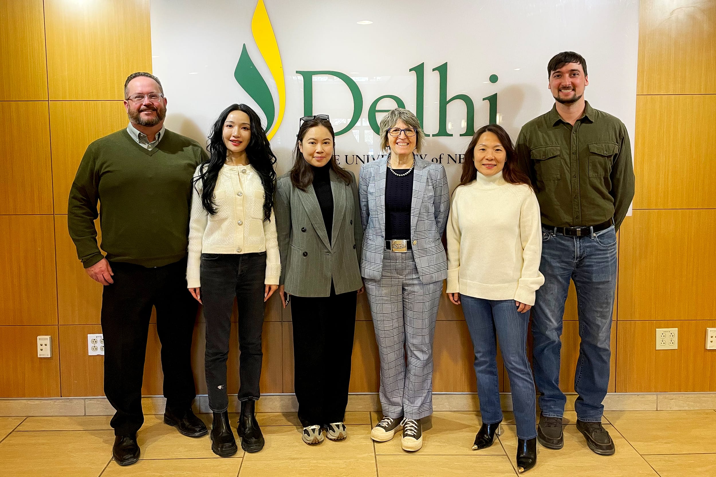 Six people stand smiling in front of a wall with the Delhi and State University of New York logo and yellow accents.