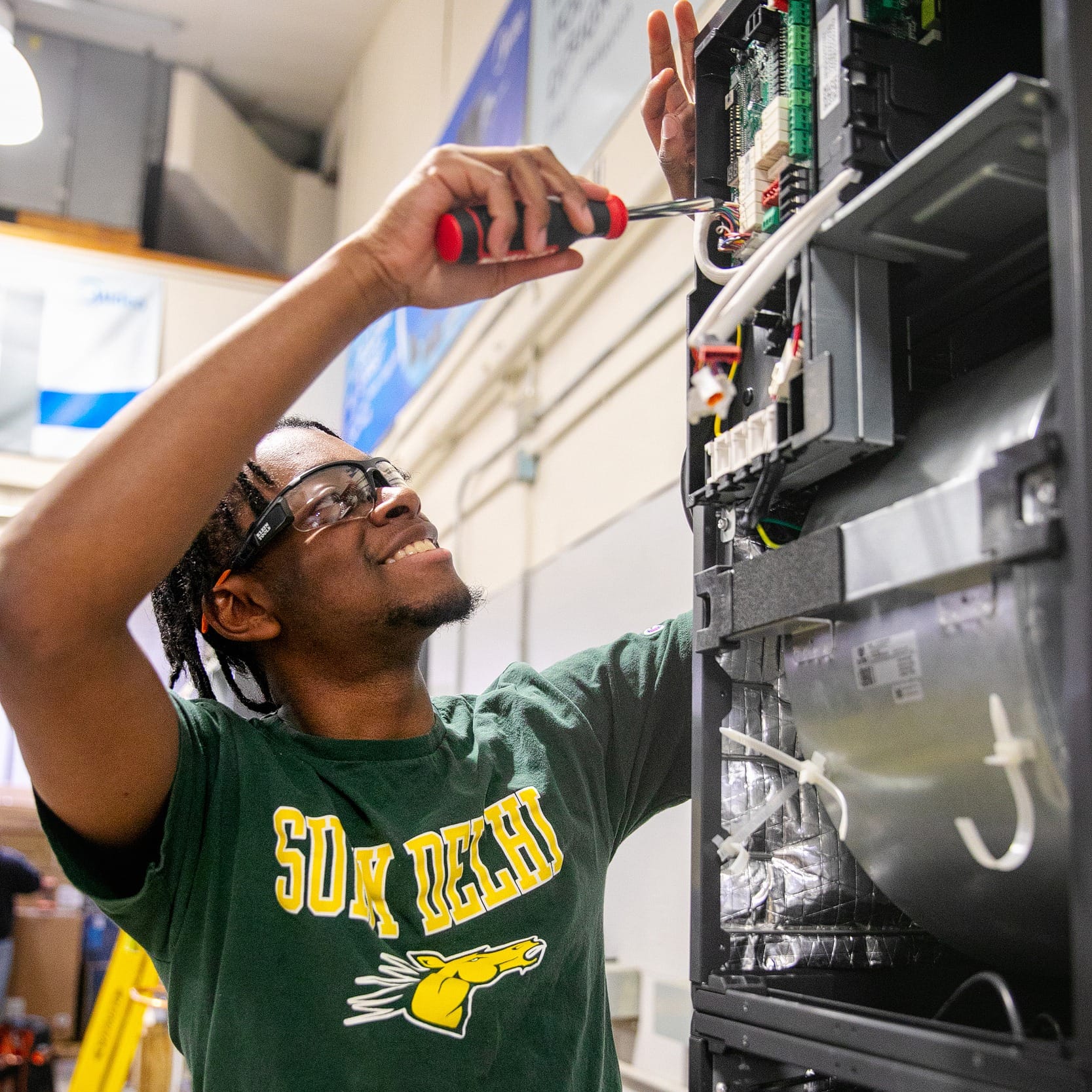 A person in a SUN DEVIL shirt uses a screwdriver to work on electronic equipment in a workshop.
