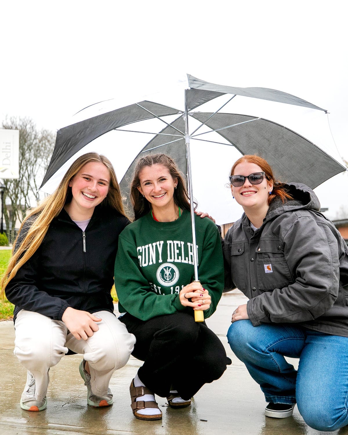 Three young women smiling outdoors under an umbrella on a rainy day, one wearing a SUNY Delhi sweatshirt.
