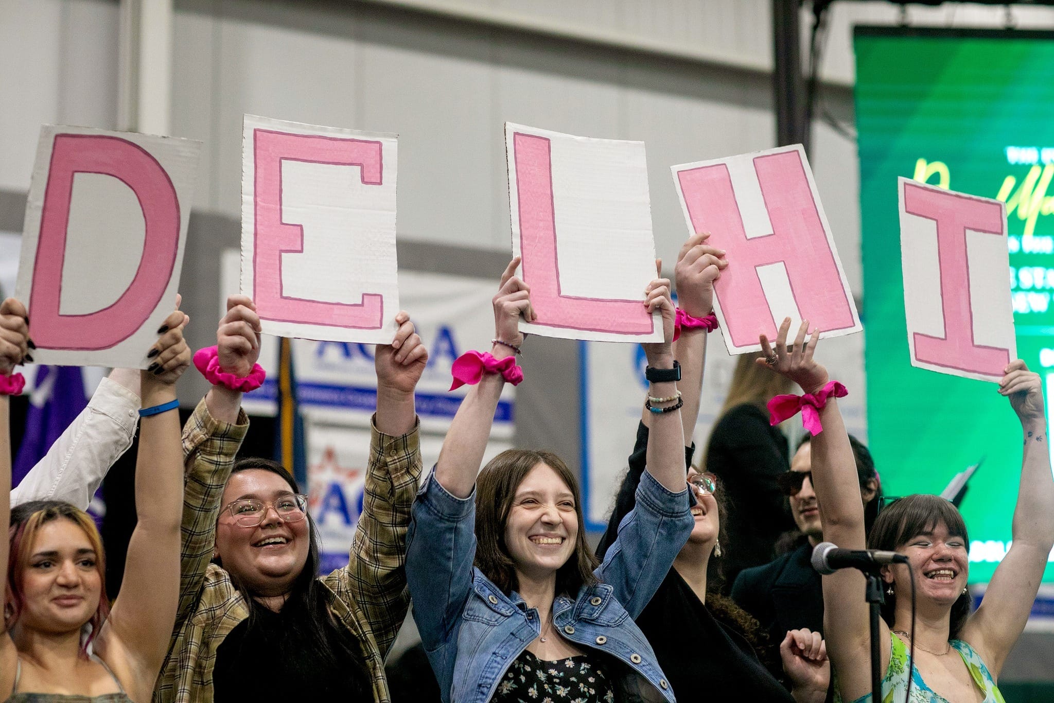 A group of smiling people hold up pink signs spelling DELHI at an indoor event.