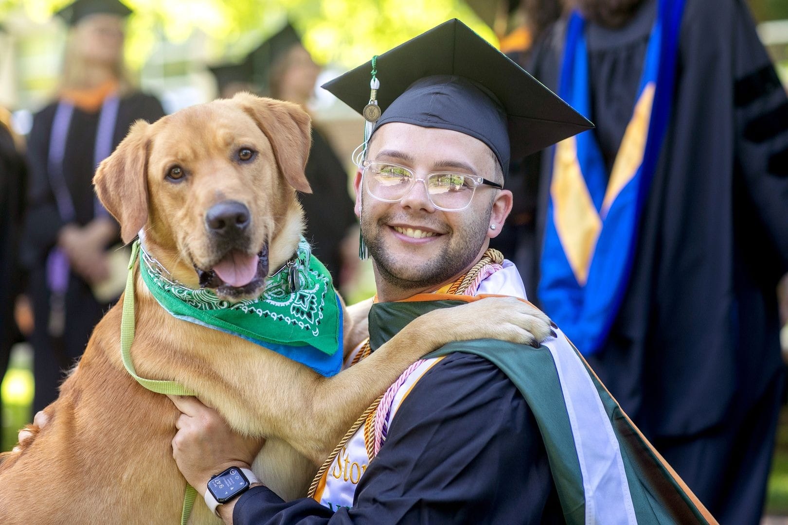 A smiling graduate in cap and gown hugs a happy dog wearing a green bandana at an outdoor graduation ceremony.