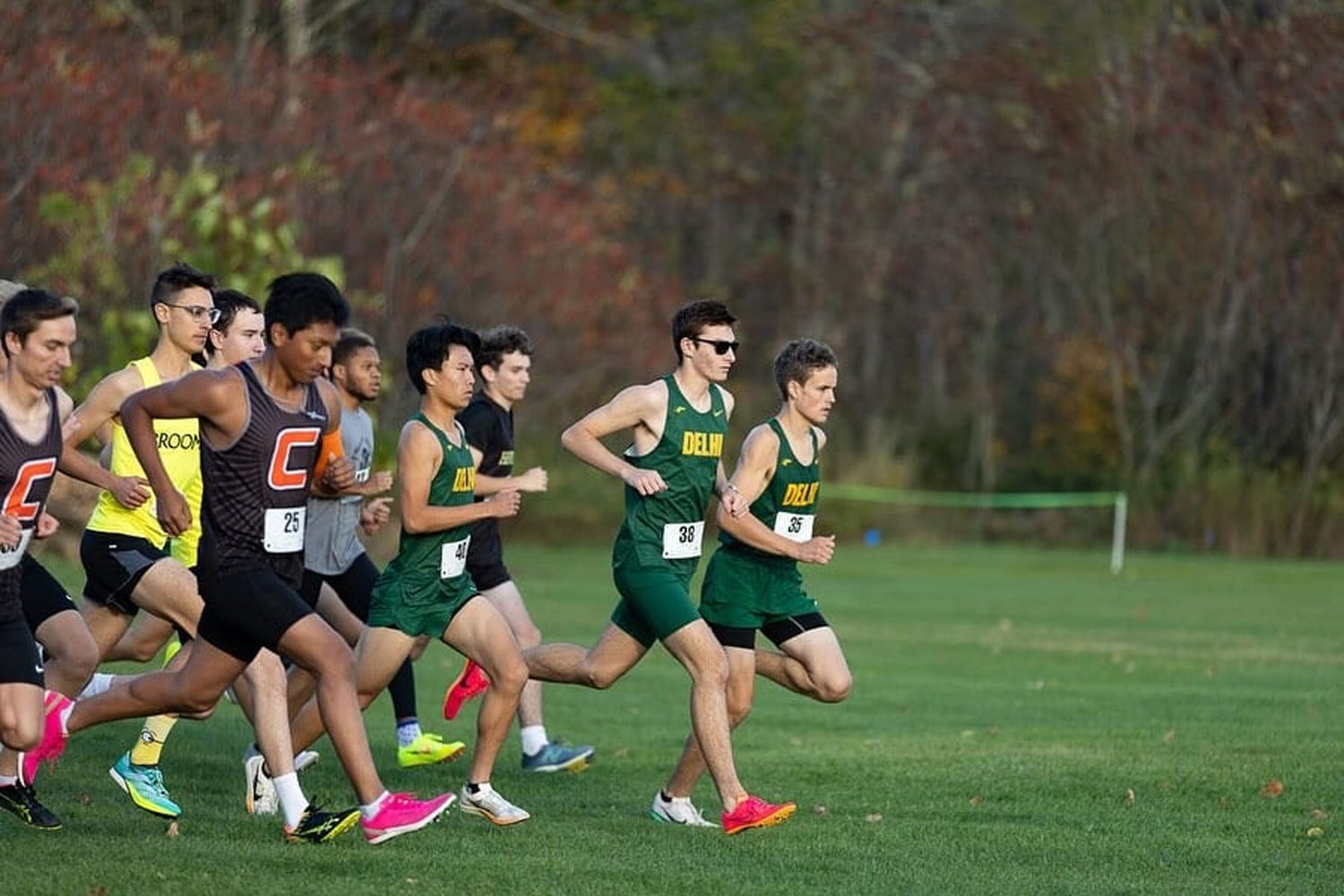 A group of male runners in athletic uniforms start a cross-country race on a grassy field.