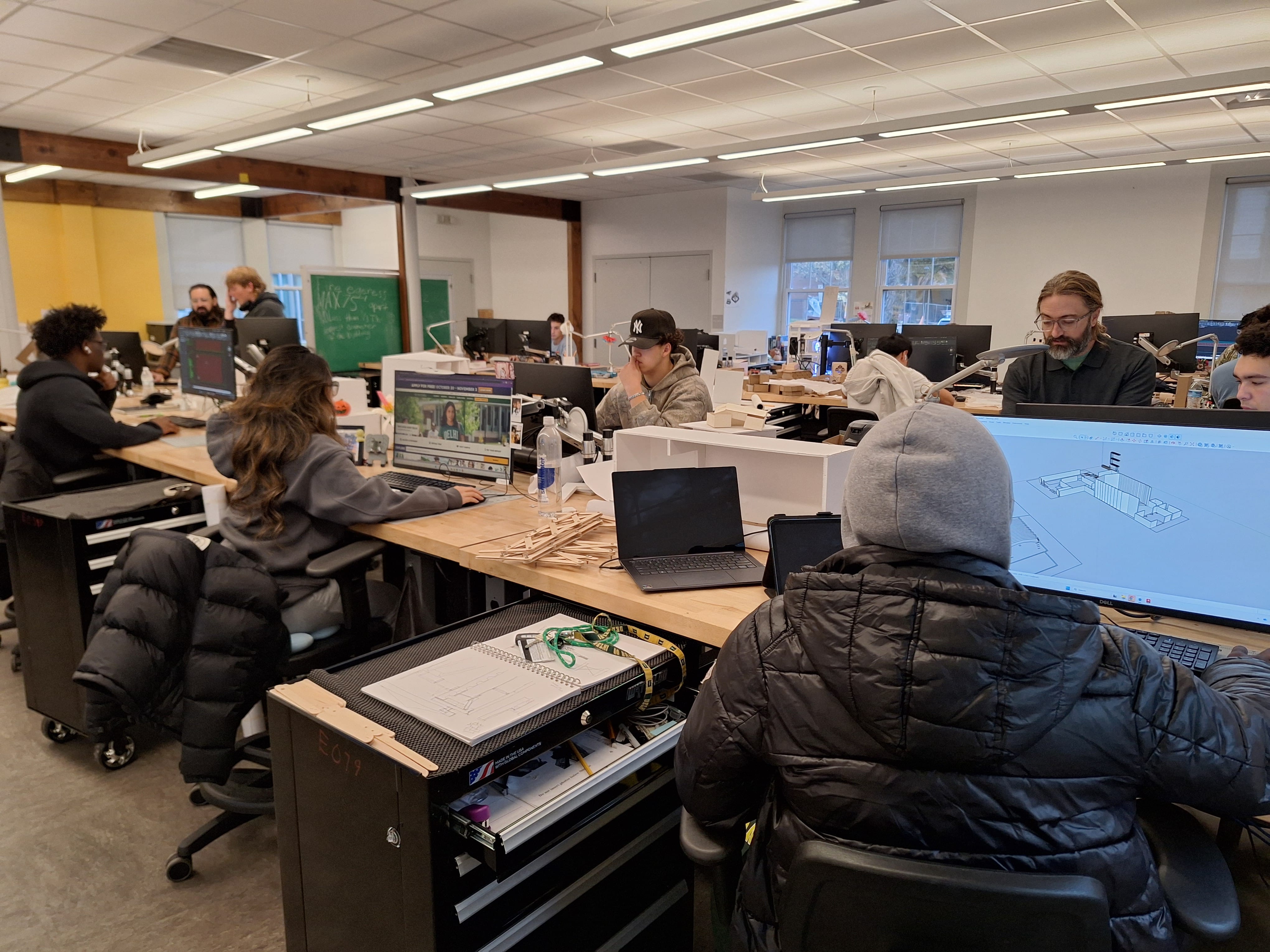 People working at desks with computers and architectural models in a bright, modern office or studio setting.