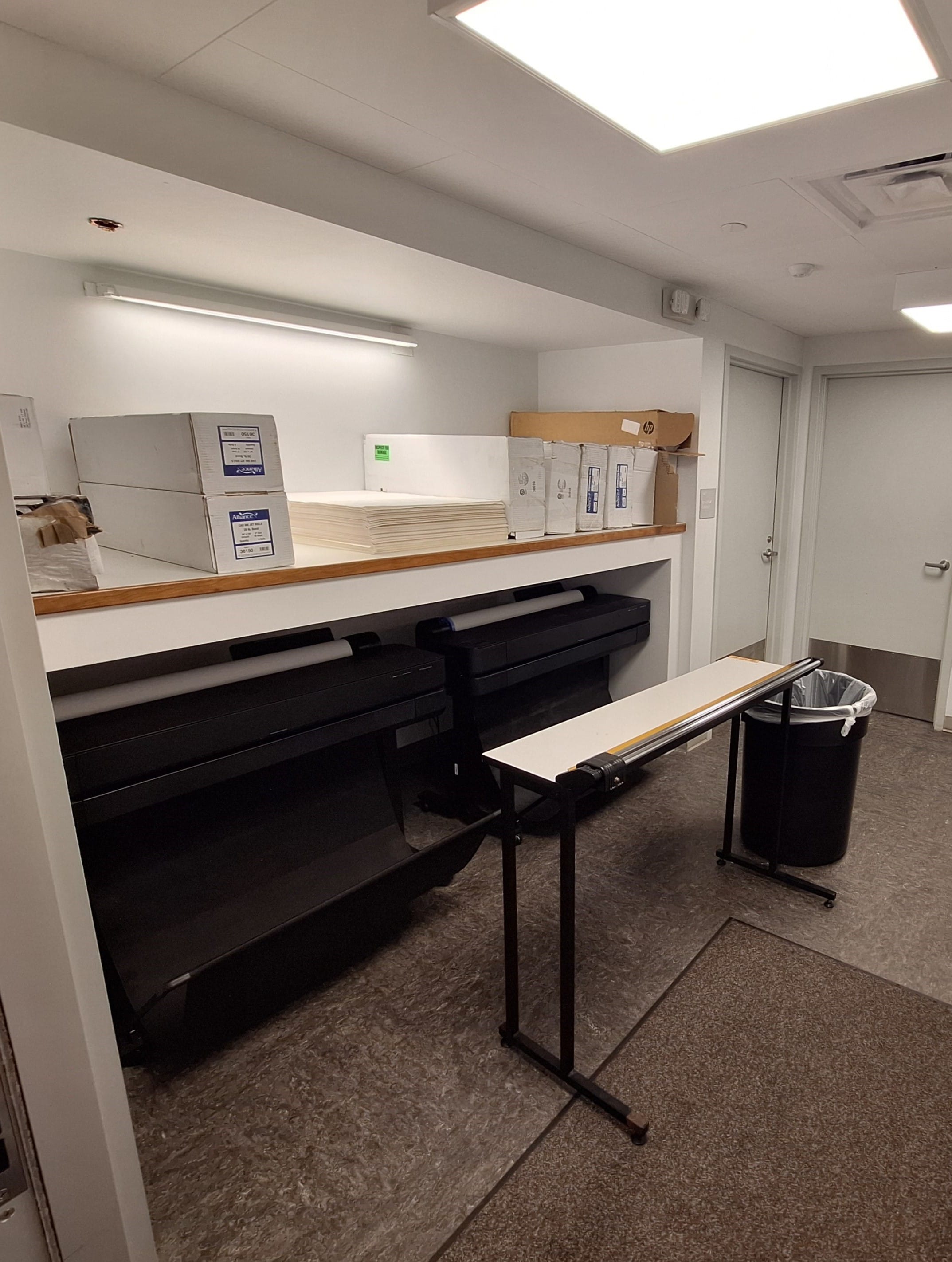 A small office room like those in drafting studios, with large printers, boxes on shelves, a table, and a trash can near the door.