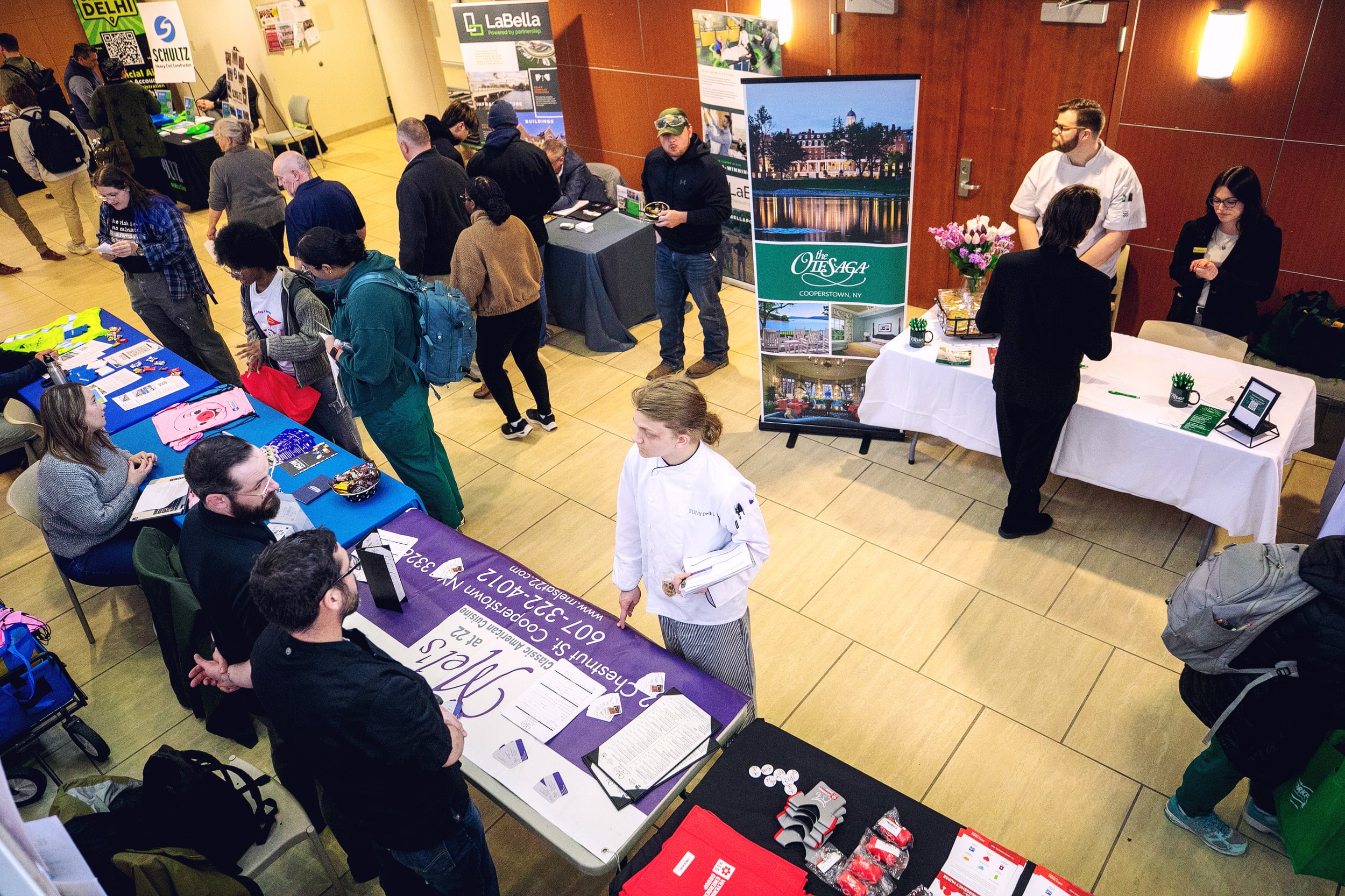 People visit various booths at an indoor career or college fair, engaging with representatives and collecting materials.